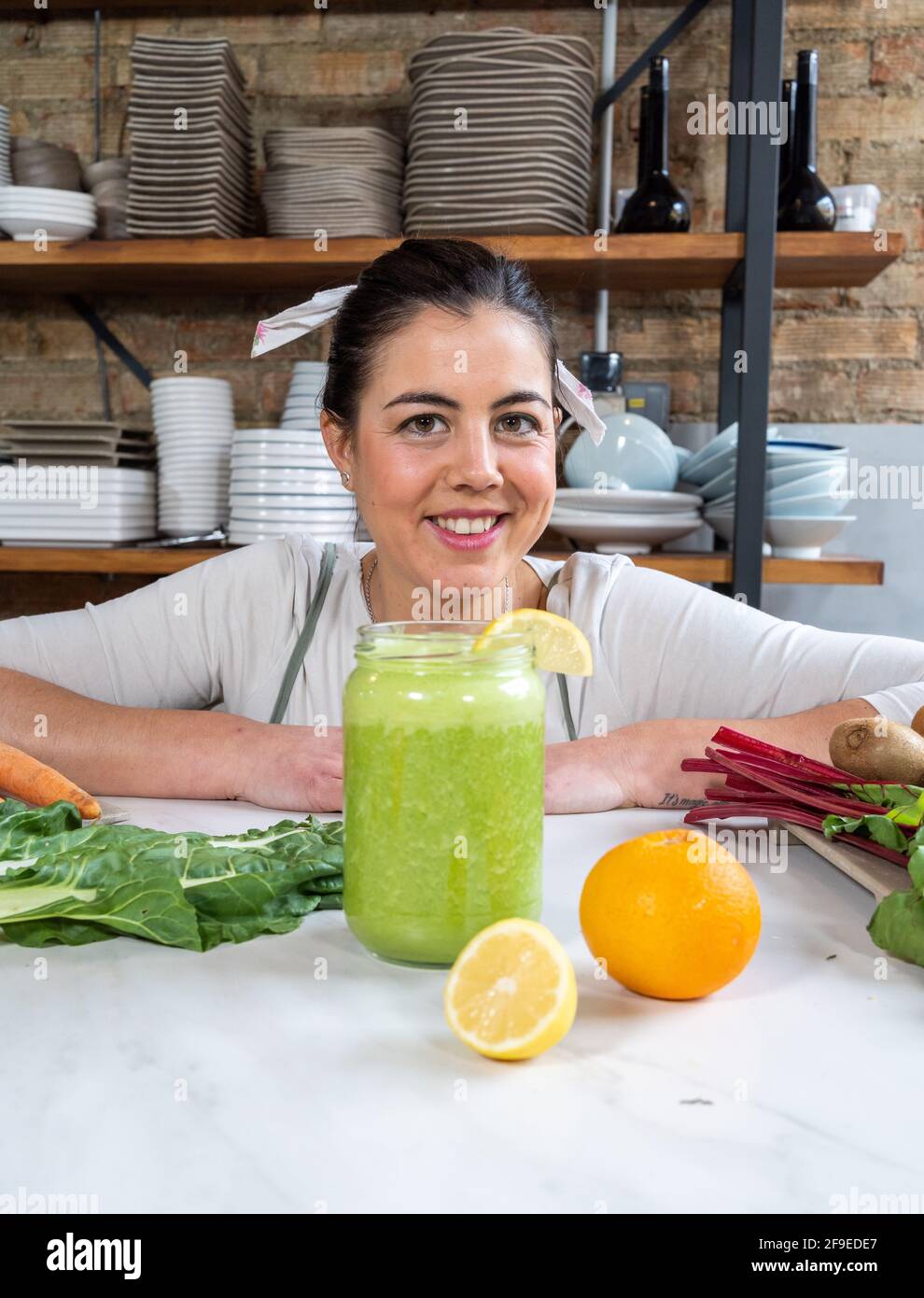 Femme joyeuse regardant l'appareil photo à la table avec un pot de délicieux smoothie végétalien et agrumes frais dans la cuisine Banque D'Images