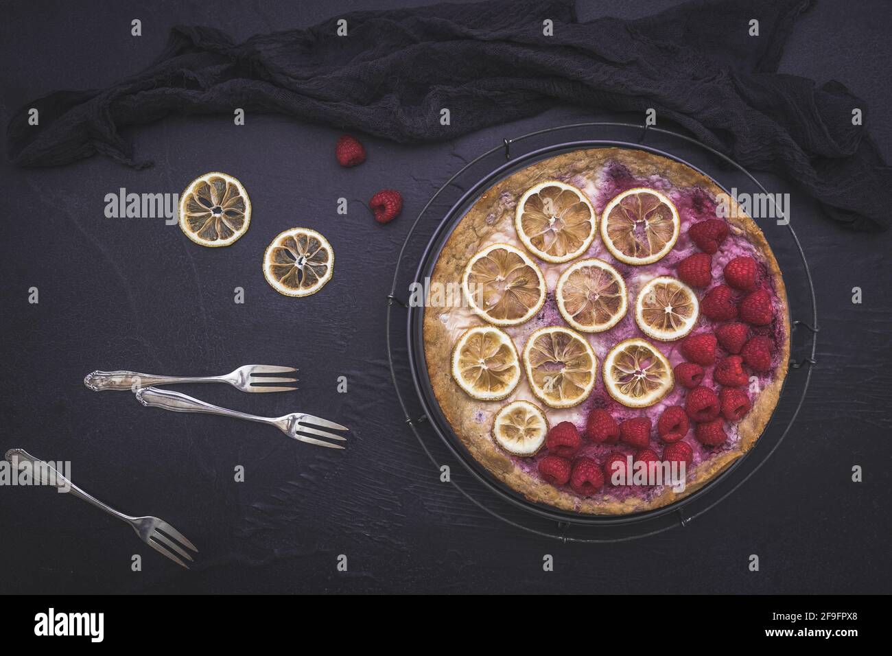 Gâteau au fromage aux framboises, tranches de citron séchées, fourchettes et tissu noir sur fond sombre, vue du dessus Banque D'Images