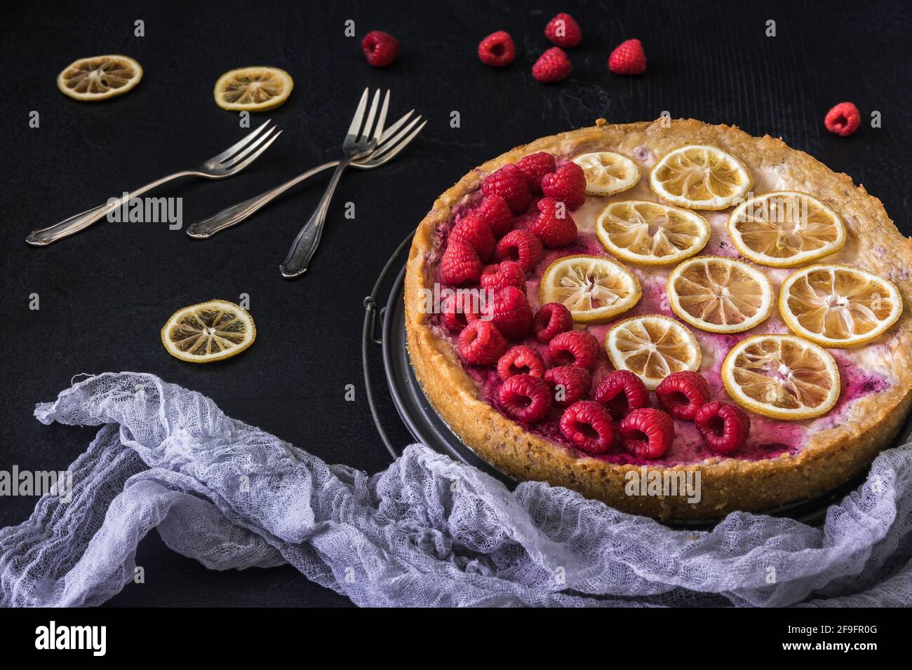 Gâteau au fromage avec framboises, tranches de citron séchées, fourchettes et toile de lilas sur fond sombre Banque D'Images