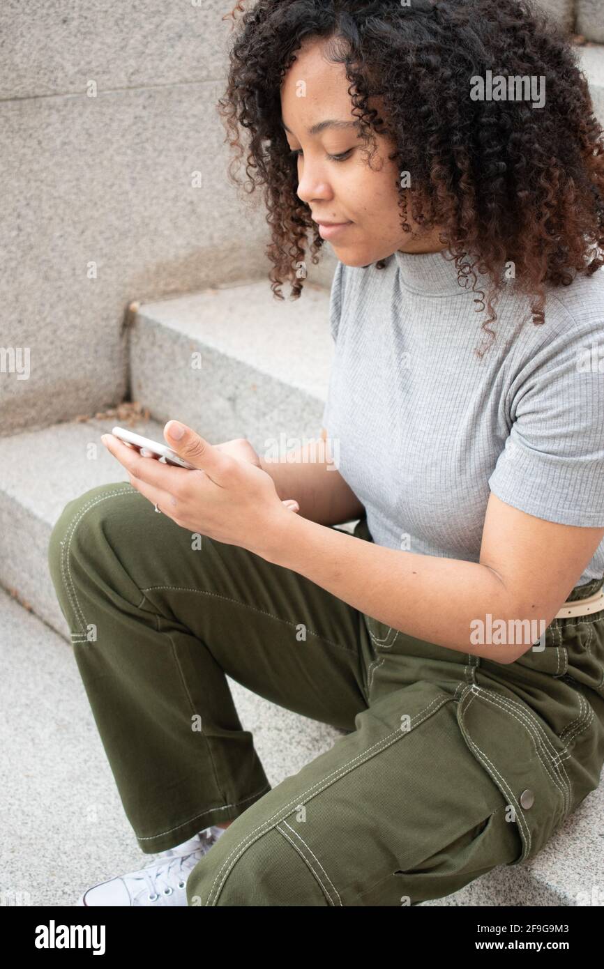 Une femme aux cheveux bouclés utilisant un téléphone portable tout en étant assise dans les escaliers. Banque D'Images