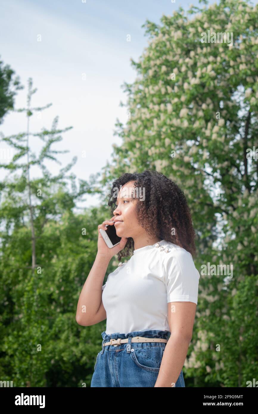 Jeune fille aux cheveux bouclés parlant sur un téléphone cellulaire tout en regardant hors du cadre. Arrière-plan des arbres verts hors foyer. Photo verticale. Banque D'Images