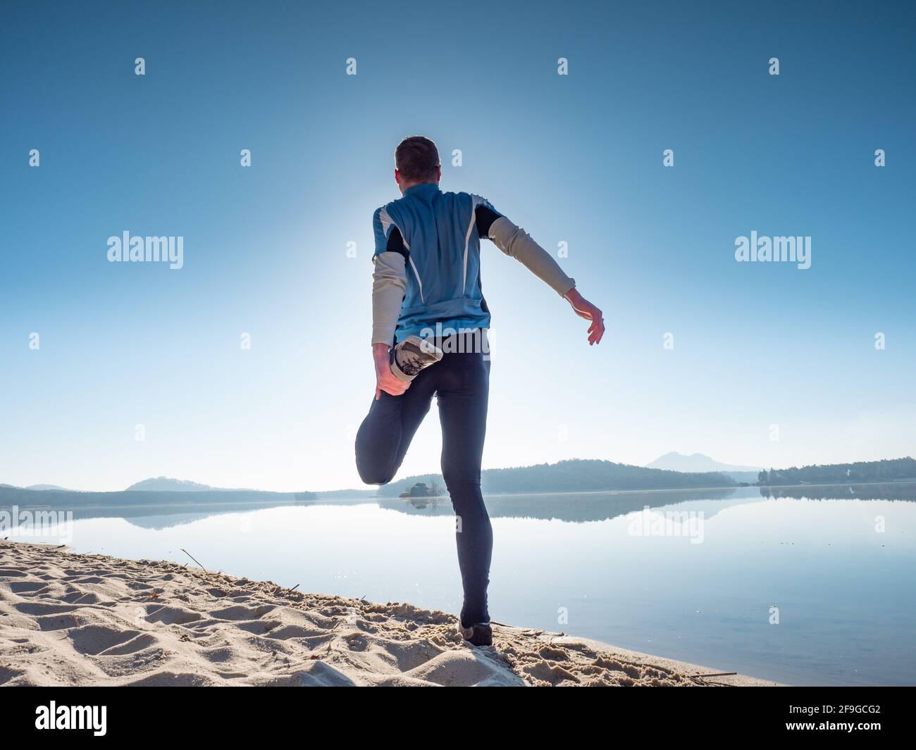 L'athlète étire les muscles de sa cuisse sur le rivage de la plage avant de courir. Entraînement matinal ensoleillé Banque D'Images