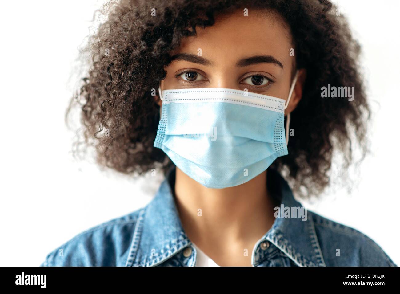 Gros plan de la jeune fille brune afro-américaine, debout à l'intérieur avec un masque médical de protection sur son visage et regarde directement dans l'appareil photo. Précautions de quarantaine, concept de soins de santé Banque D'Images