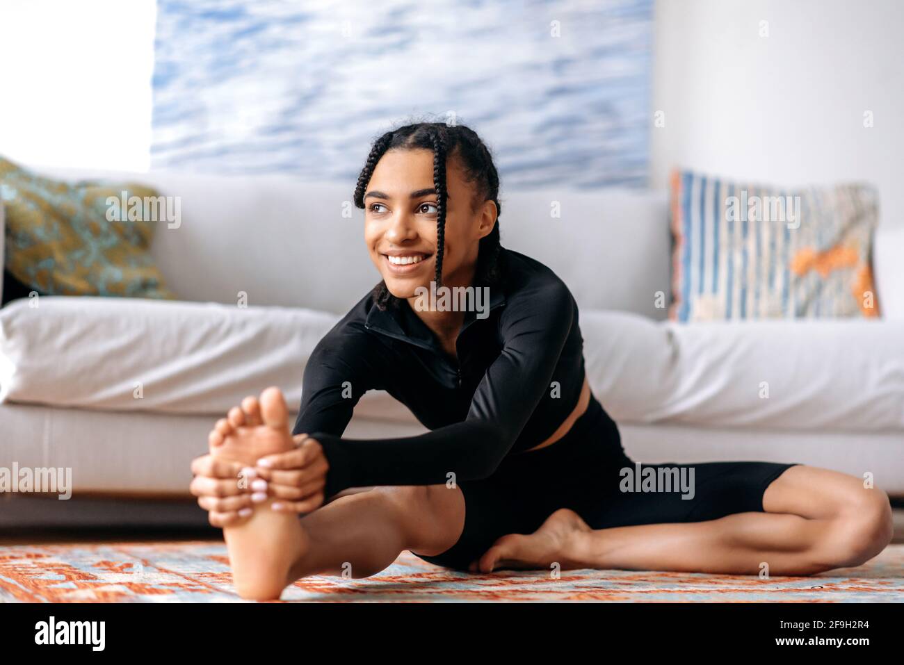 Sportive et flexible jolie fille afro-américaine vêtue de vêtements de sport, est engagé fitness, mène un style de vie sain, ne s'étirant à la maison sur le sol, regarde sur le côté, sourires heureux Banque D'Images