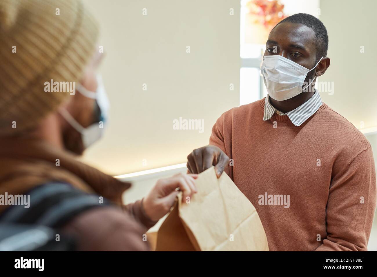 Portrait de l'homme afro-américain qui distribue l'ordre de emporter au client dans un café, tous deux portant des masques, concept de restrictions de covid Banque D'Images