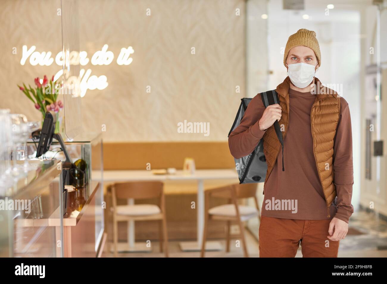 Vue avant portrait de l'homme de livraison portant un masque et regardant l'appareil photo tout en se tenant à l'intérieur du café, espace de copie Banque D'Images