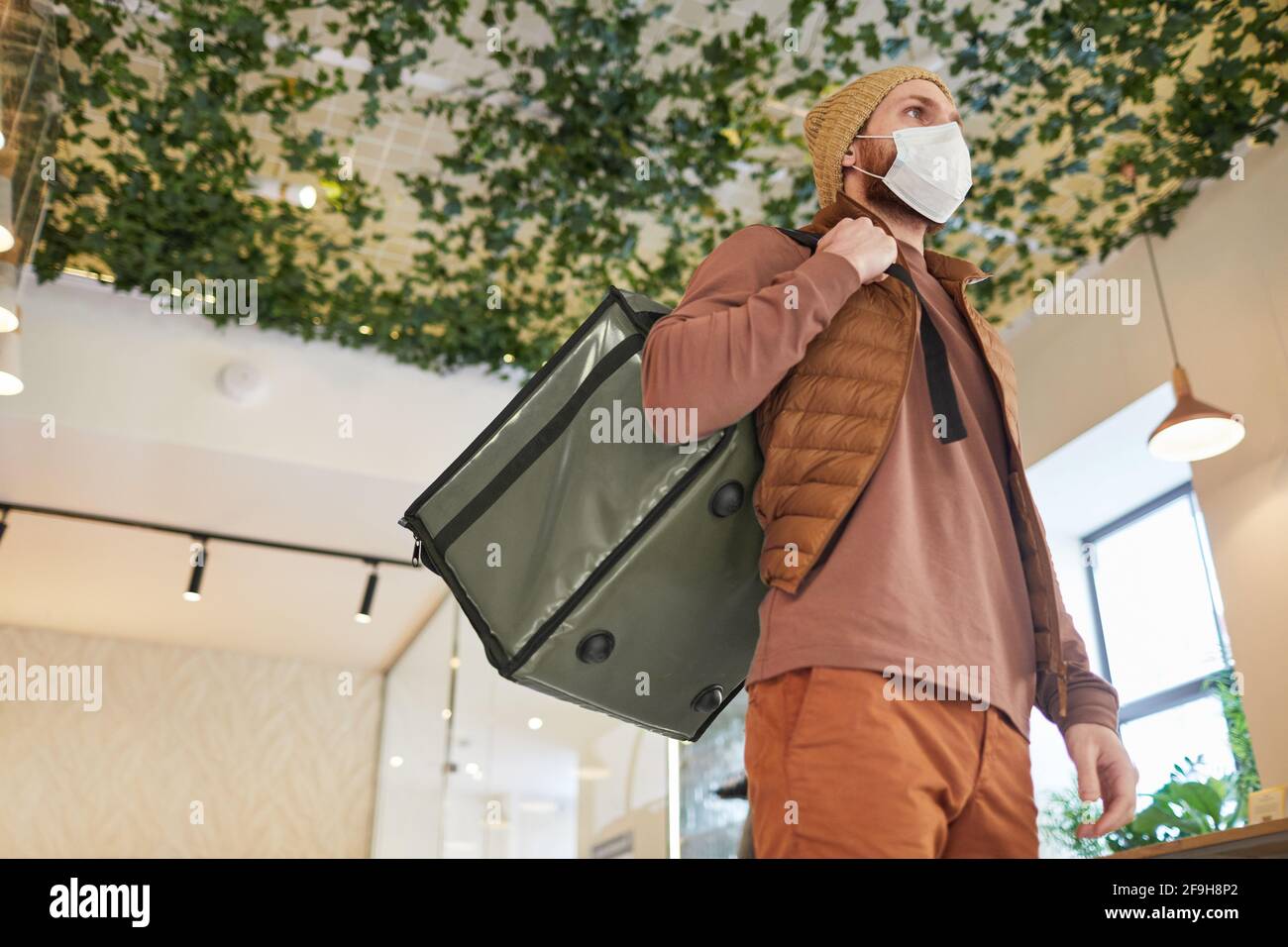 Portrait à angle bas de l'homme de livraison portant un masque et tenant le sac tout en se tenant à l'intérieur du café, espace de copie Banque D'Images