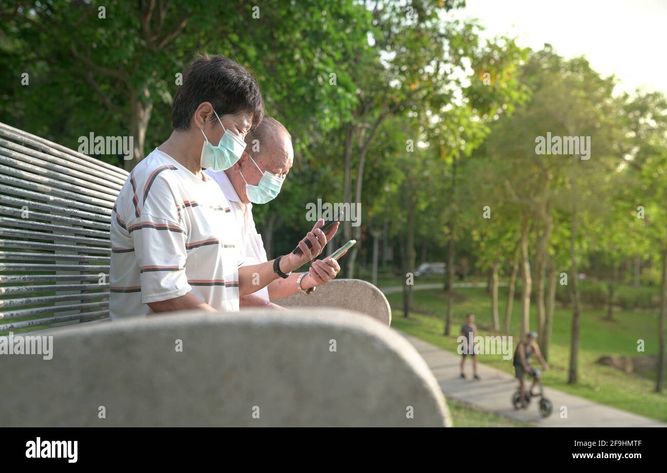 Couple asiatique senior portant un masque, assis et lisant leur téléphone dans un parc. Banque D'Images