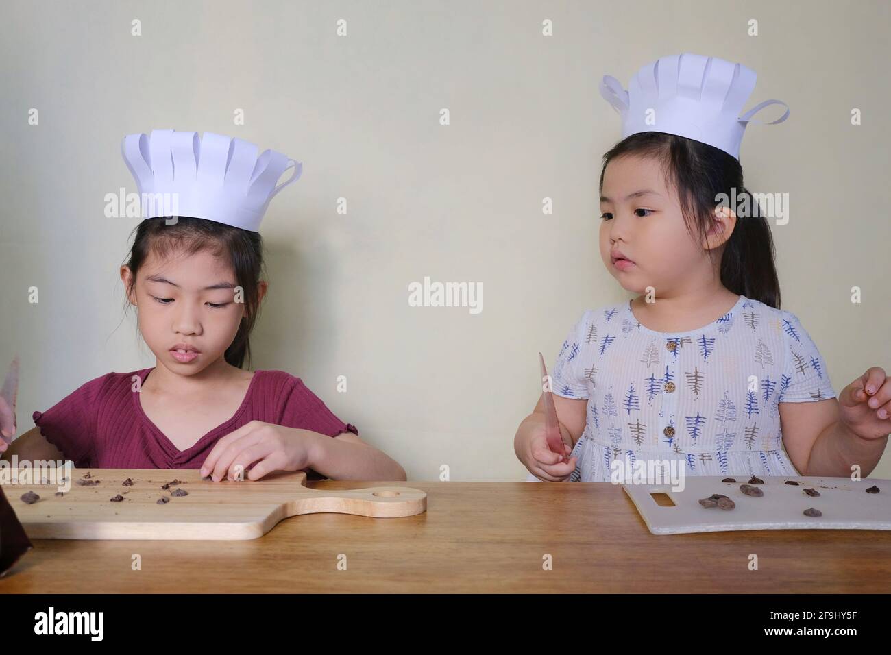 Deux jeunes sœurs asiatiques mignonnes, avec de jolis chapeaux de chef, apprennent à fabriquer des biscuits aux pépites de chocolat, en coupant du chocolat en petits morceaux avant de mixer Banque D'Images