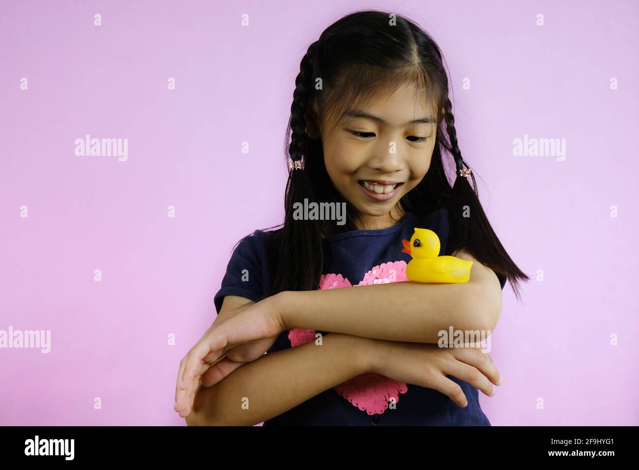 Une jeune fille asiatique mignonne joue avec joie avec son petit canard en caoutchouc jaune, l'équilibrant sur son coude avec ses bras croisés, souriant et amusant. Banque D'Images