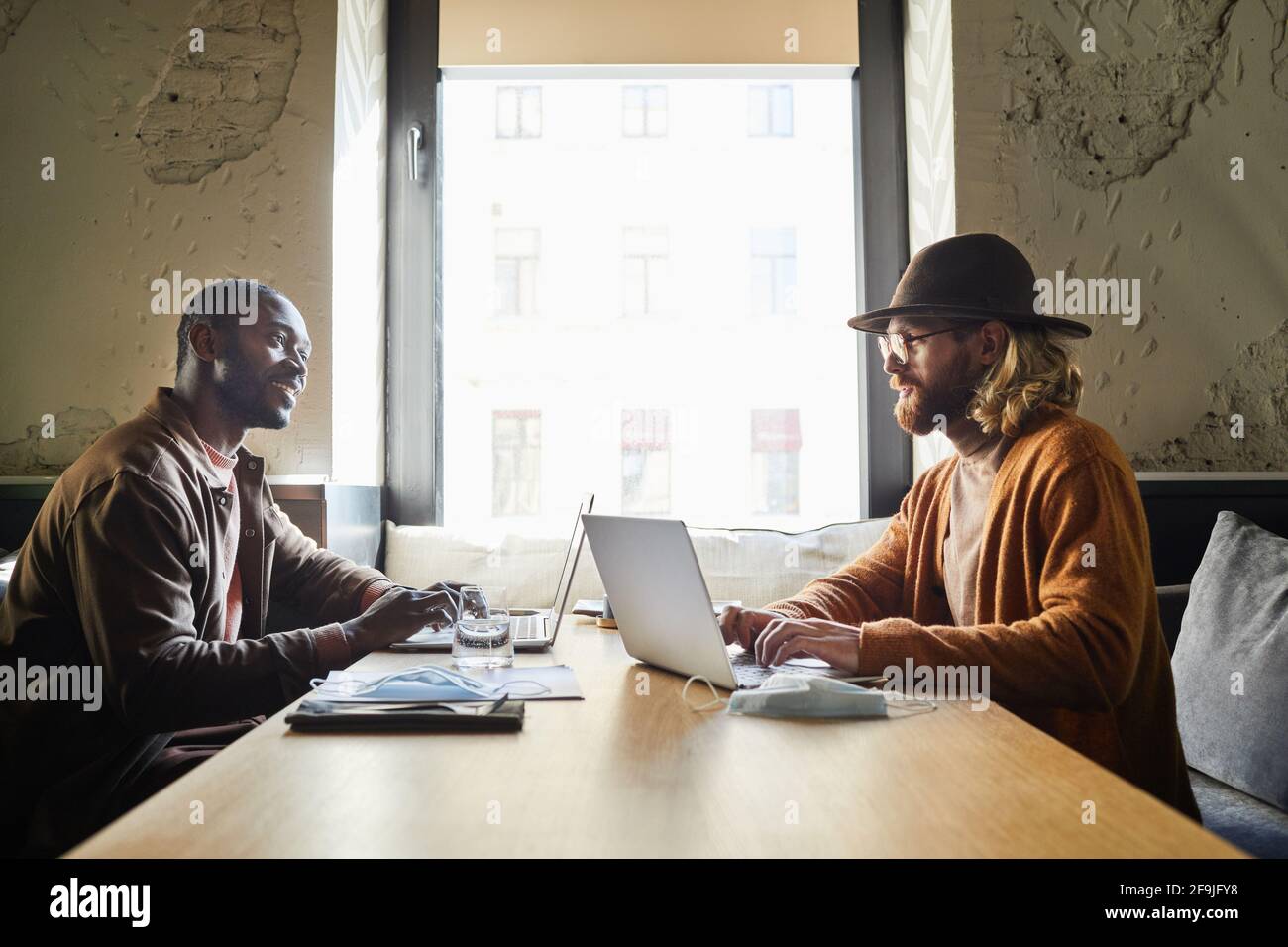 Vue latérale de deux hommes d'affaires souriants utilisant un ordinateur portable tout en travaillant à une table de café éclairée par la lumière du soleil dans l'intérieur du loft, espace de copie Banque D'Images