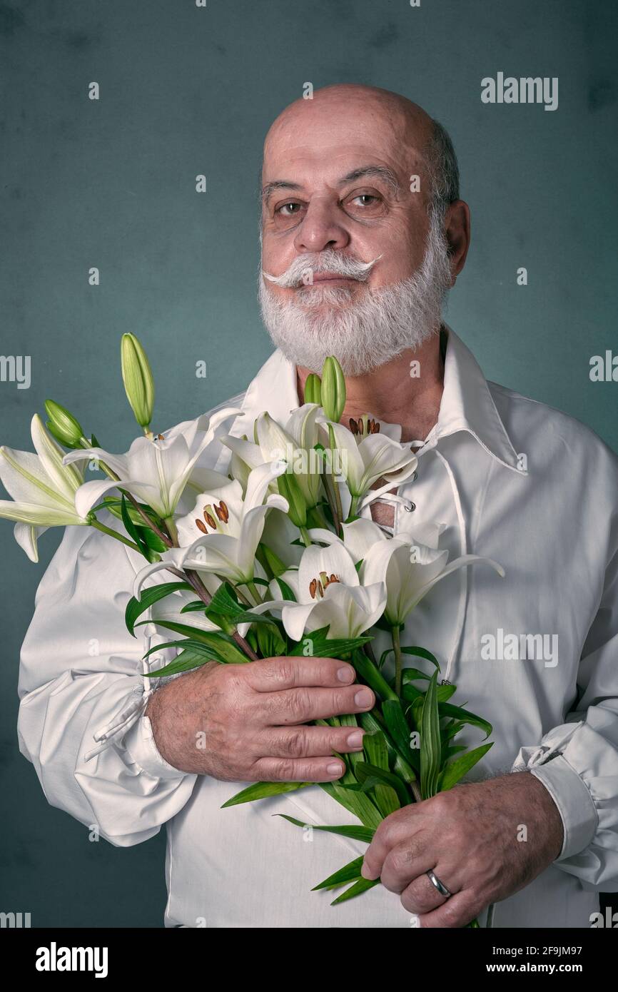 Portrait d'un homme âgé, chauve, avec une barbe grise et d'apparence latine, portant une chemise blanche et tenant un bouquet de lys blancs Banque D'Images