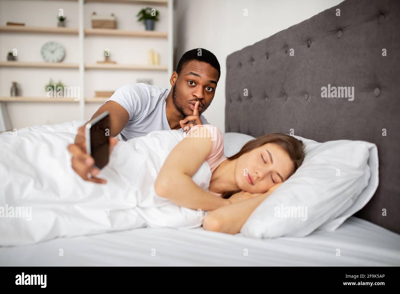 Un mari noir jaloux espionne sa femme, montrant un geste DE RUSE, vérifiant son téléphone mobile pendant qu'elle est endormi au lit Banque D'Images