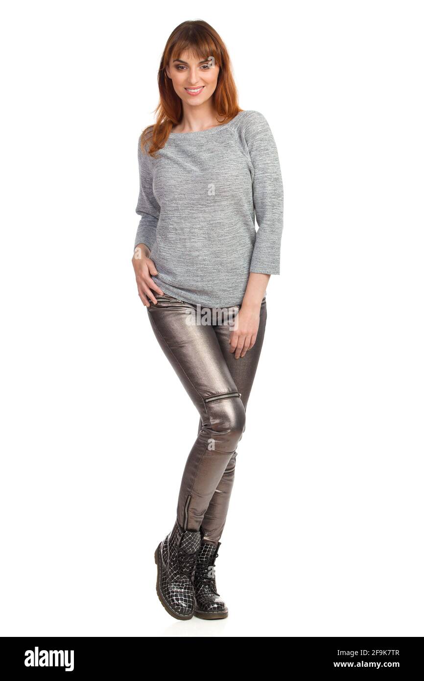 Jeune femme en blouse grise, pantalon brillant et bottes noires est debout et souriant. Vue avant. Prise de vue en studio sur toute la longueur isolée sur blanc. Banque D'Images