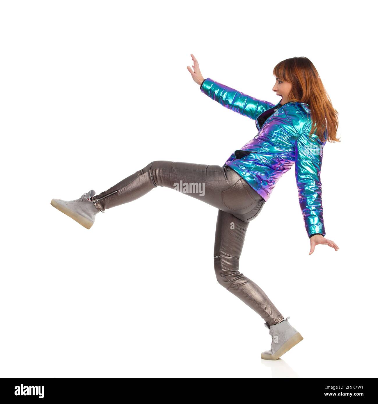 Drôle de jeune femme en veste de duvet, pantalon brillant et baskets est en train de faire un grand pas et crier. Vue latérale. Prise de vue en studio sur toute la longueur isolée sur W Banque D'Images