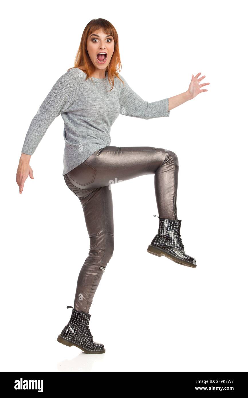 La jeune femme açée dans un chemisier gris, un pantalon brillant et des bottes noires marche, regarde l'appareil photo et crie. Vue latérale. Prise de vue en studio pleine longueur isolée Banque D'Images