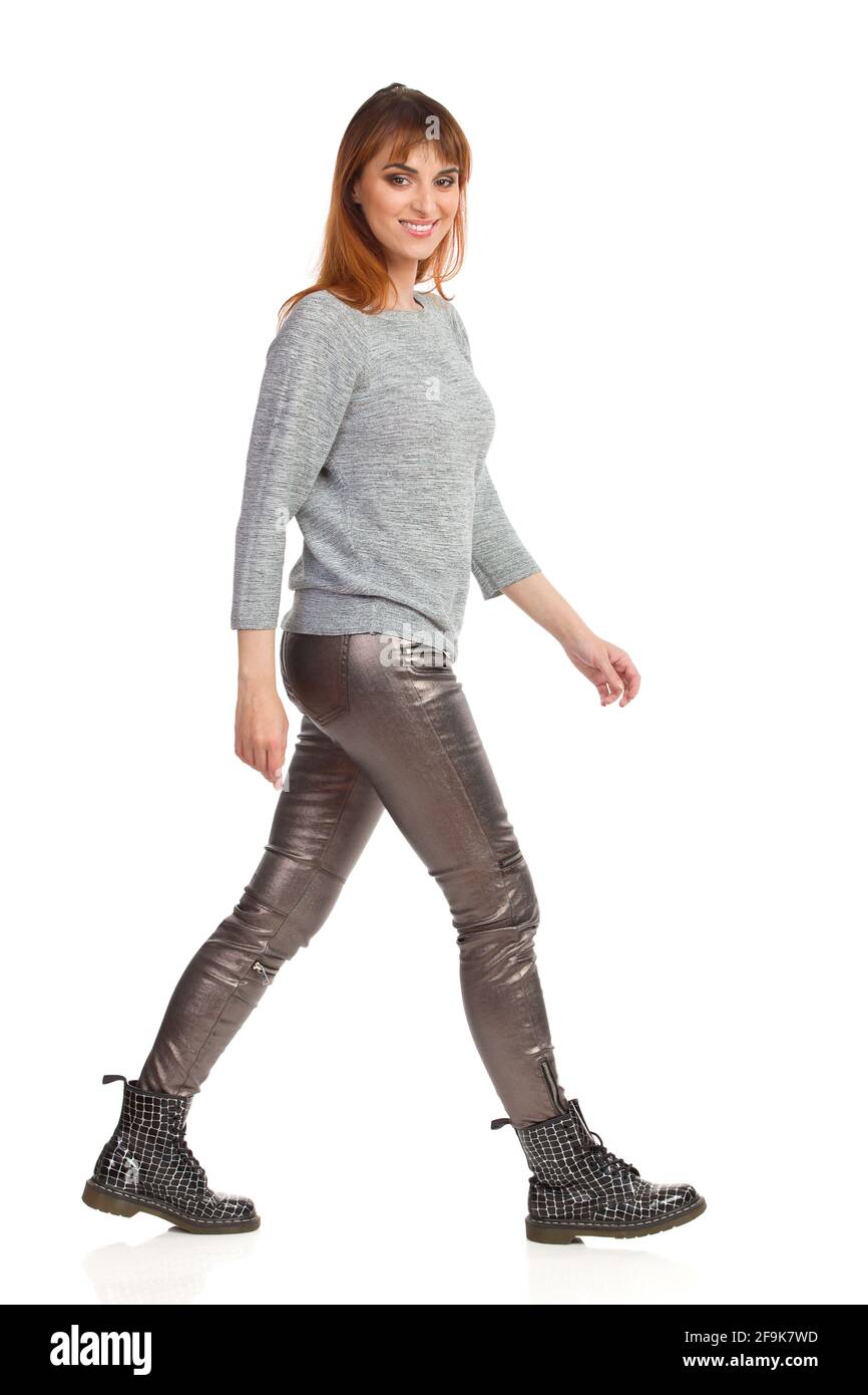 Une jeune femme en blouse grise, pantalon brillant et bottes noires marche, regarde l'appareil photo et sourit. Vue latérale. Prise de vue en studio sur toute la longueur isolée sur le coup Banque D'Images