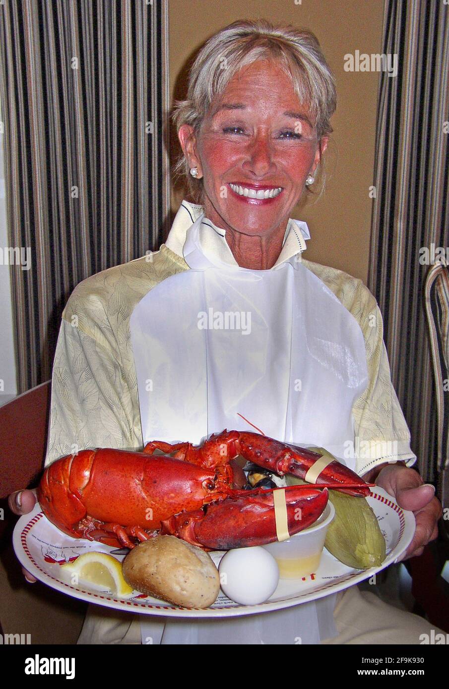 Une femme au dîner présente avec joie un homard américain (Homarus americanus) qui domine l’assiette en papier qui contient un repas populaire appelé « ébullition du homard ». La nourriture comprend les grands crustacés et les plats d'accompagnement qui comprennent souvent du maïs sur l'épi (ici dans sa husse), des pommes de terre à peau rouge, des œufs durs, des petits pains et des palourdes, ainsi que du beurre fondu dans un petit récipient et des tranches de citron. Situé le long de la côte nord de l'Amérique du Nord, ce délicieux poisson de mer est connu sous le nom de Maine, canadien, Atlantique, Nord et vrai homard. Normalement de couleur brun-verdâtre, ils deviennent rouges lorsqu'ils sont bouillis Banque D'Images