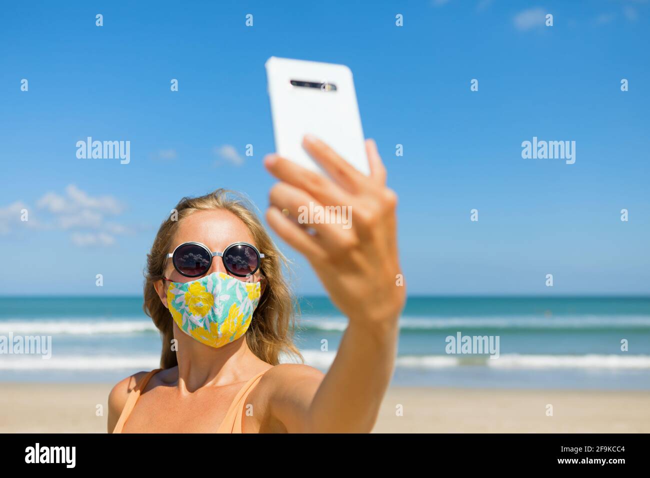 Drôle de fille prenant photo selfie par smartphone sur la plage tropicale de mer. Nouvelles règles pour porter un masque de couverture en tissu dans les lieux publics en raison de coronavirus COVID Banque D'Images