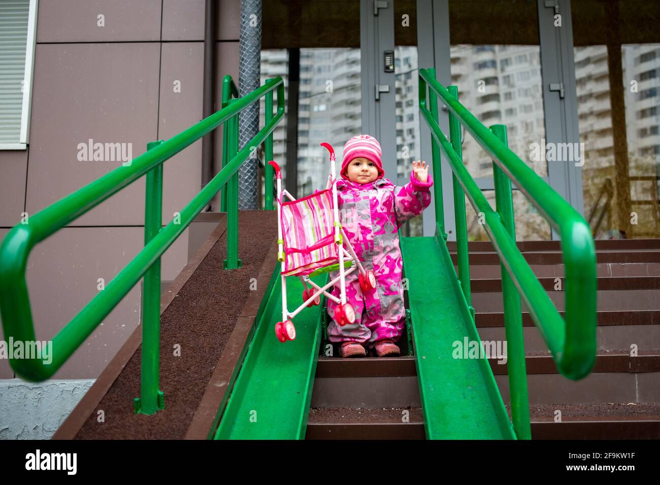 un adorable tout-petit avec une poussette en peluche marche le long d'une rampe  de garde-corps en acier pour fauteuil roulant, chariots et poussettes.  descente douce des escaliers à l'extérieur Photo Stock -