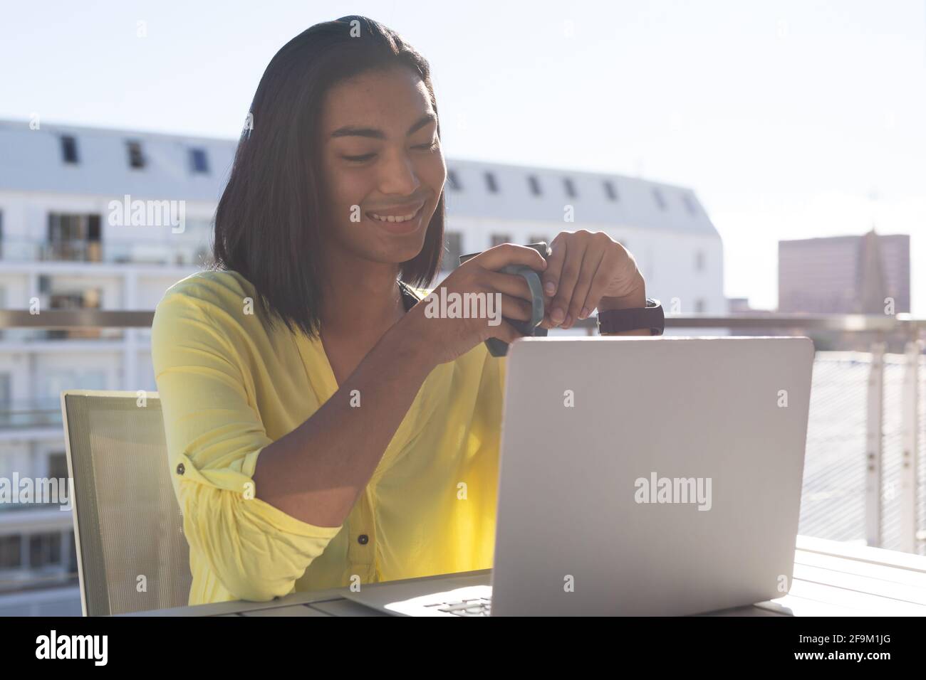 Femme transgenre souriante et mixte assise à table sur une terrasse ensoleillée utilisation d'un ordinateur portable pour le café Banque D'Images