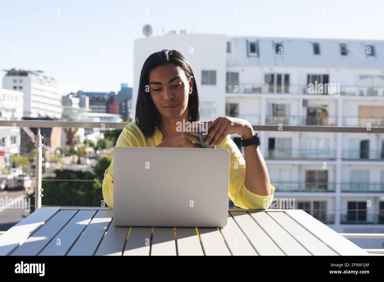 Femme souriante de race mixte transgenre assis à table sous le soleil terrasse avec ordinateur portable et café Banque D'Images