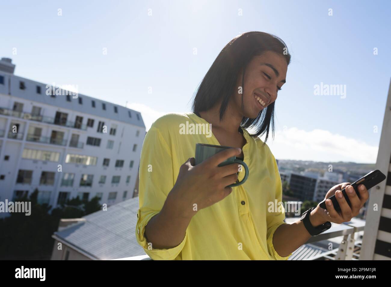 Femme souriante de race mixte transgenre debout sur la terrasse ensoleillée du toit tenir le café à l'aide d'un smartphone Banque D'Images