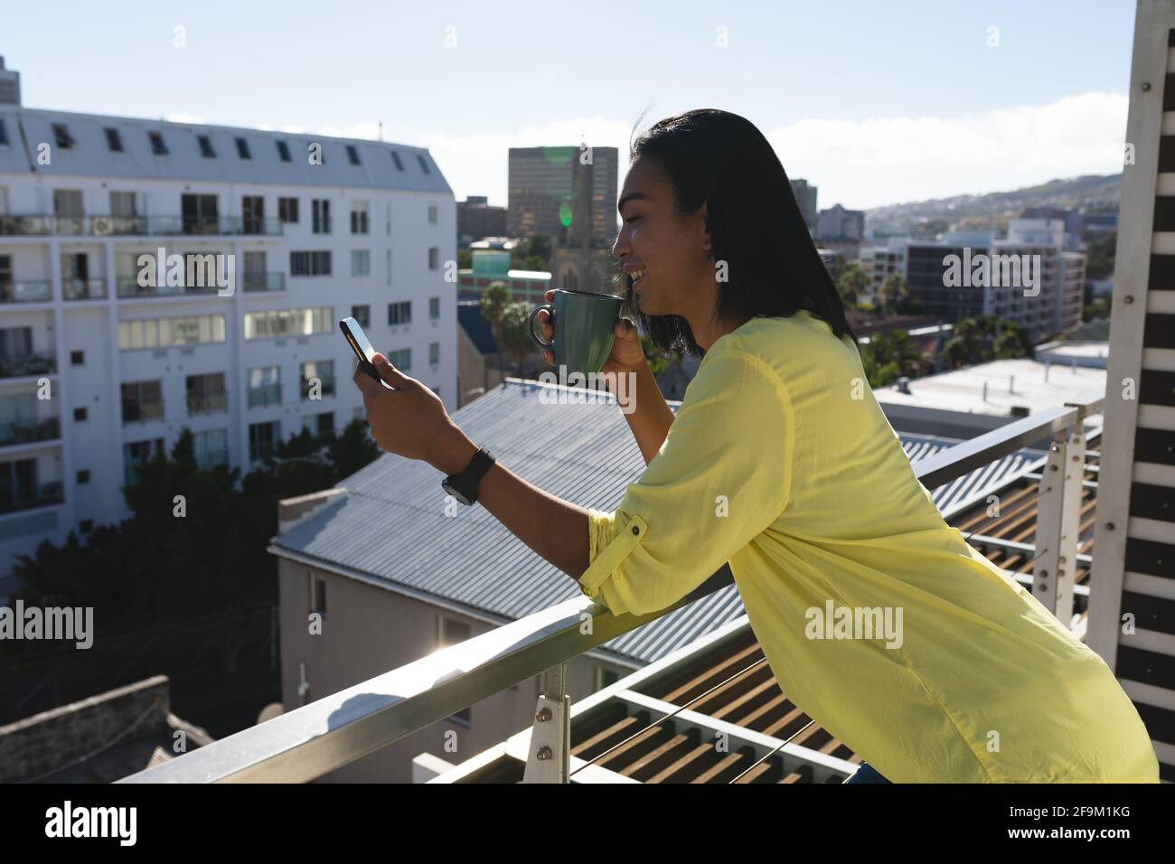 Souriant mixte transgenre femme homme debout sur la terrasse ensoleillée de toit tenir le café à l'aide d'un smartphone Banque D'Images