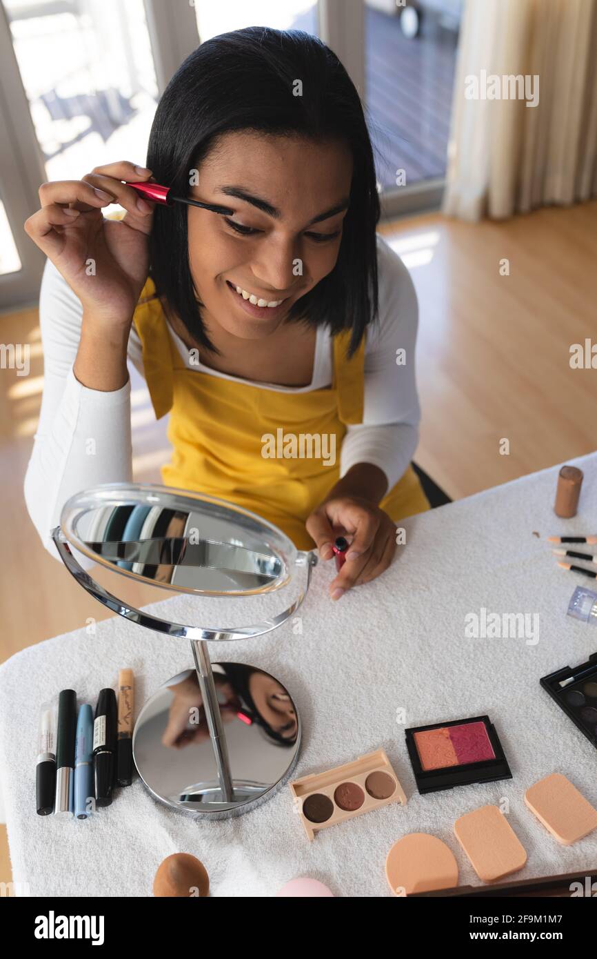 Femme souriante de race mixte transgenre assis à table regardant à l'intérieur miroir mettant sur mascara Banque D'Images