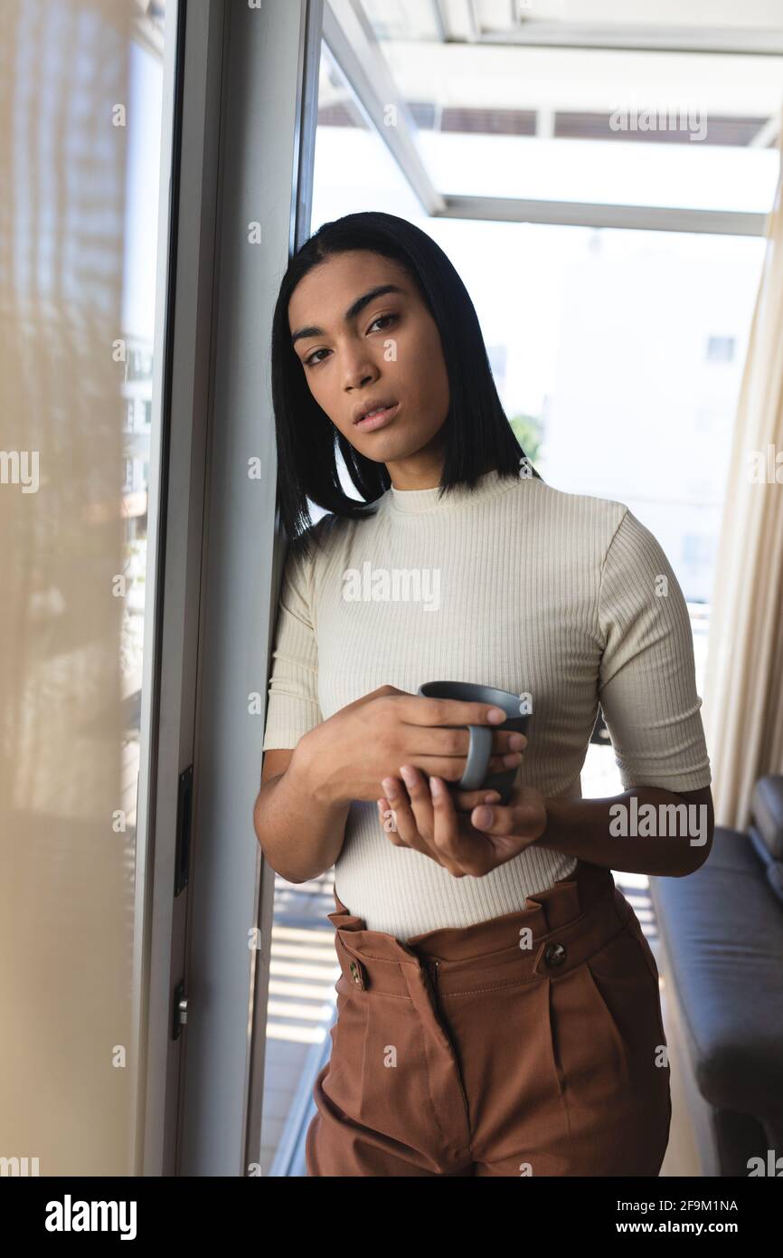 Portrait de la femme transgenre de race mixte debout par fenêtre tenue tasse de café Banque D'Images