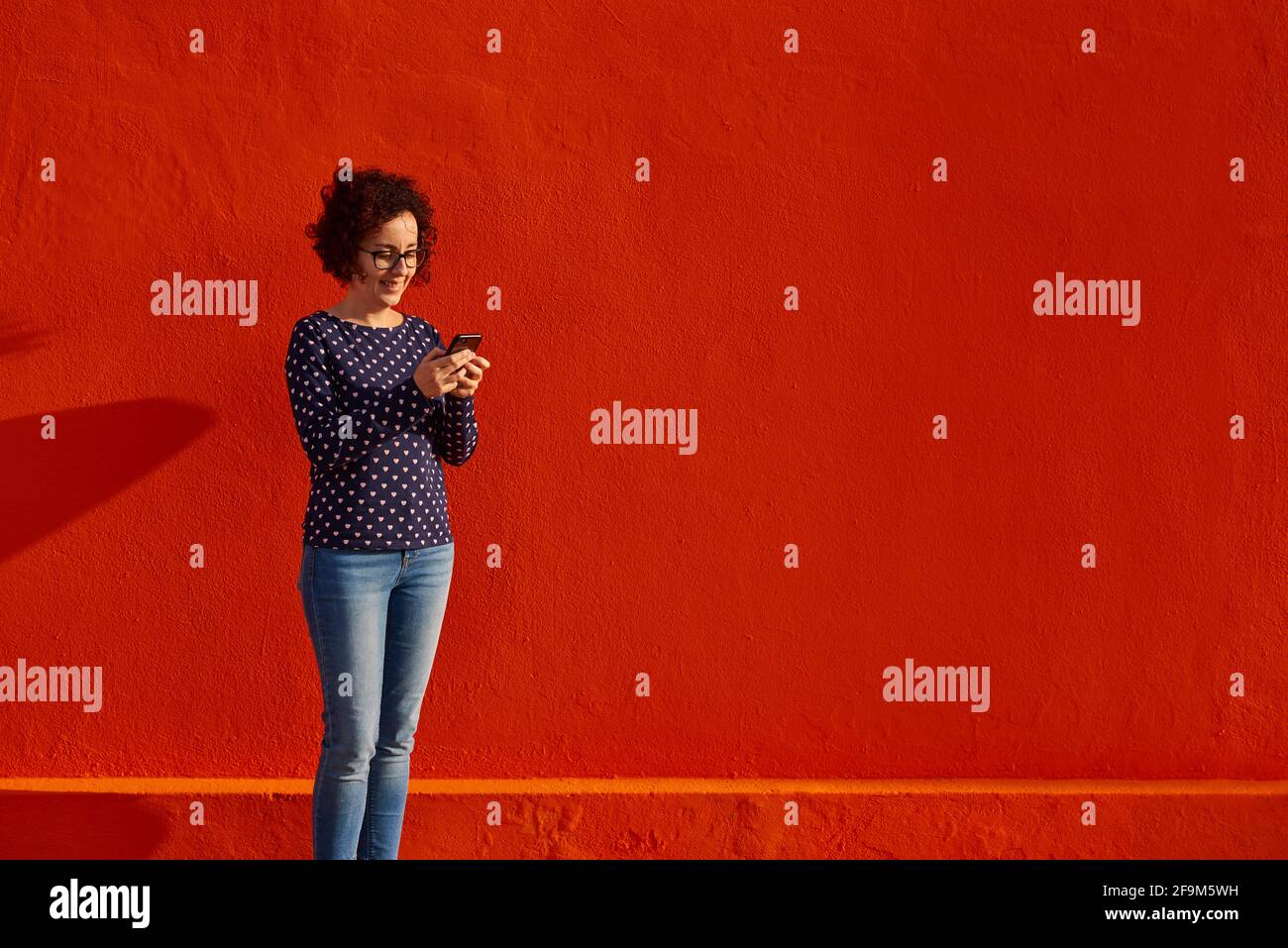 Vue avant d'une femme heureuse debout contre un mur rouge vif à l'aide de son smartphone. Photo de haute qualité. Banque D'Images