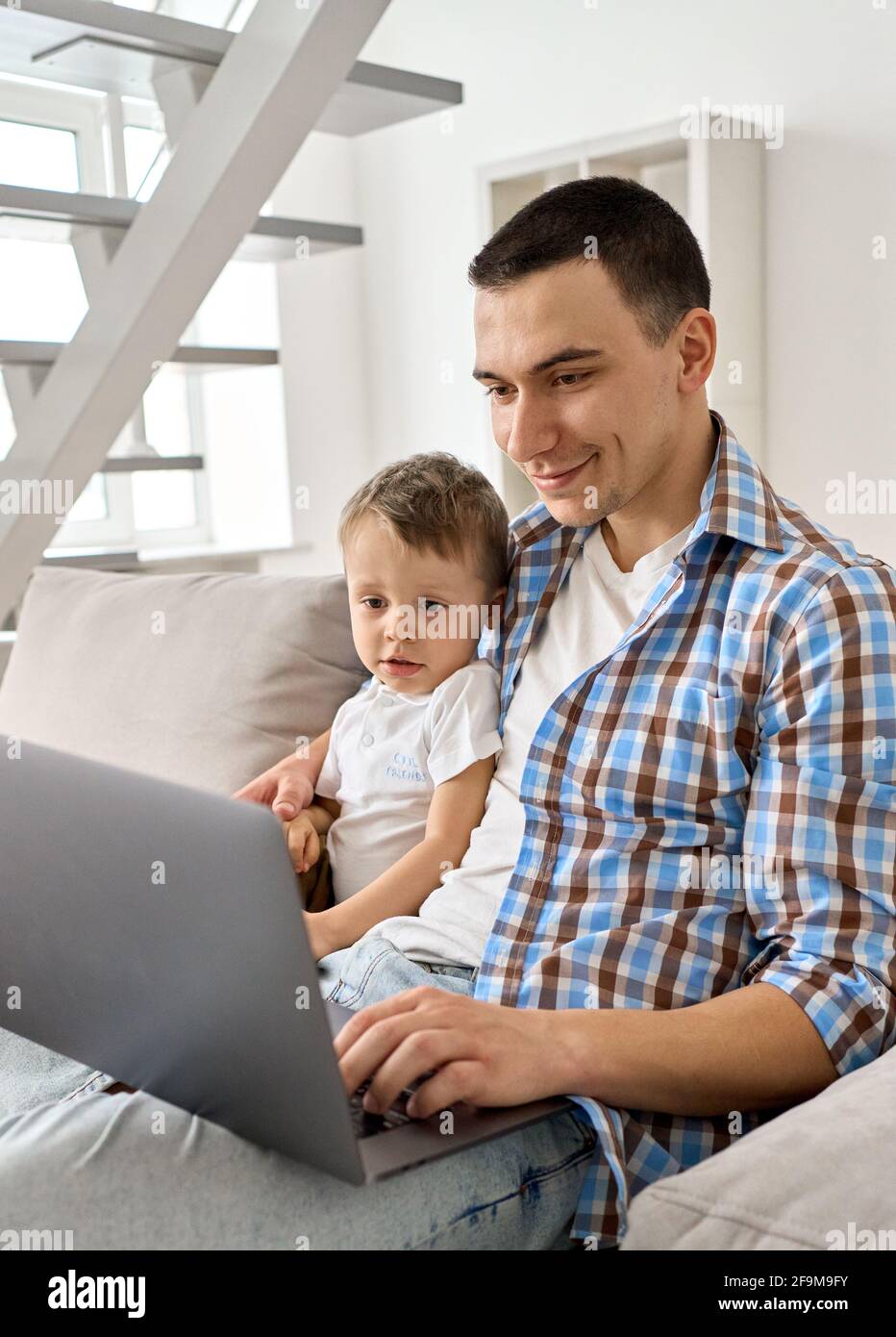Un jeune père heureux qui se moque d'un petit fils regarde un ordinateur portable assis sur un canapé. Banque D'Images