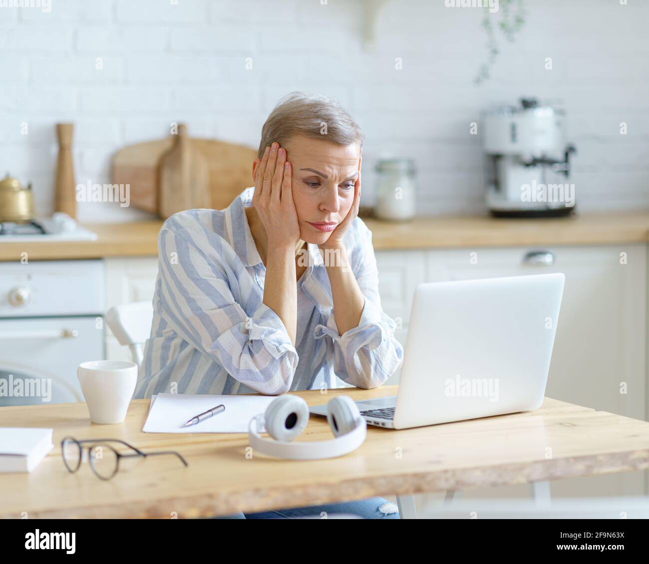 Femme mûre stressée en tenue décontractée assise dans une cuisine moderne, tenant la tête dans les mains et regardant sur l'écran d'ordinateur portable tout en travaillant ou étudiant en ligne de la maison. Concept de technologies et de personnes âgées Banque D'Images
