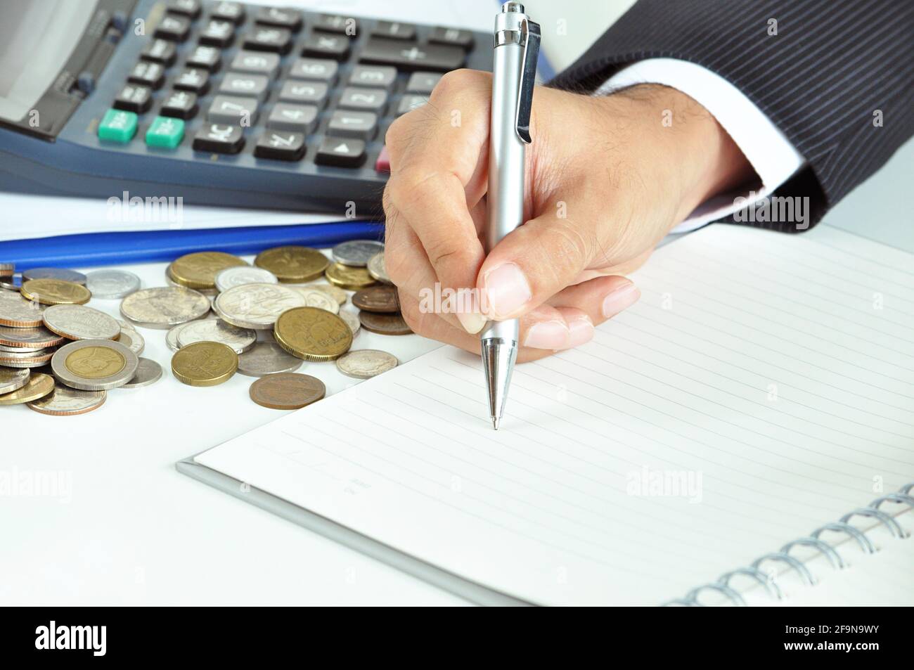 Main d'homme d'affaires tenant un stylo écrivant sur du papier vide avec pièces de monnaie et calculatrice - concept commercial et financier Banque D'Images