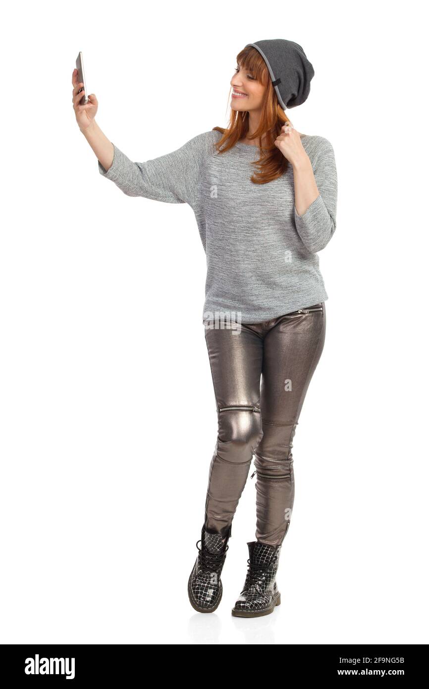 Jeune femme décontractée en blouse grise, pantalon brillant, bottes noires et casquette d'hiver tient le téléphone, parle un selfie et sourire. Prise de vue en studio Banque D'Images