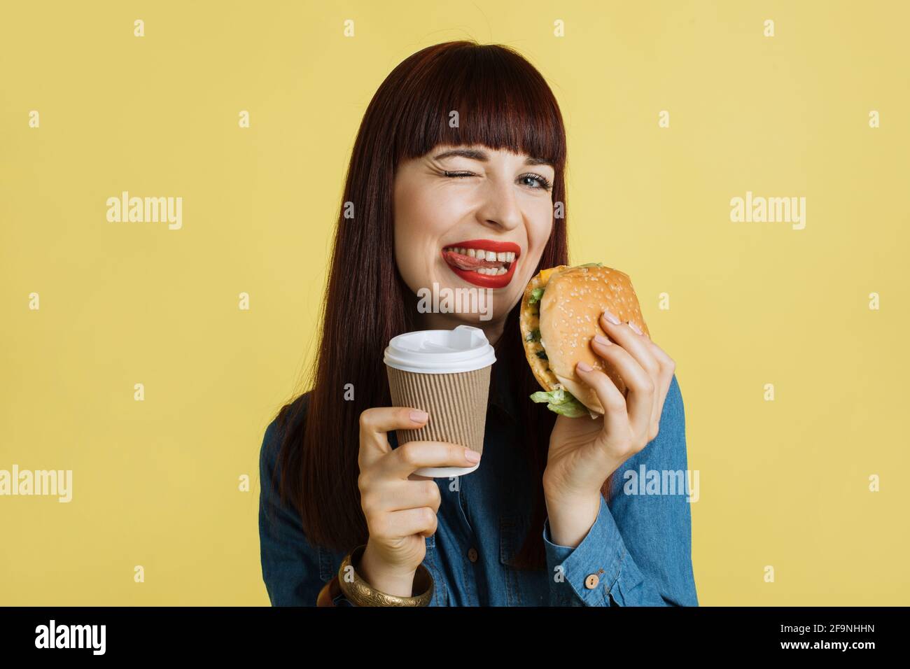 Gros plan sur une jeune femme attrayante et gaie qui grimace à l'appareil photo, montre la langue, tient un savoureux hamburger et prend le café et profite de son heure de déjeuner. Isolé sur fond de studio jaune Banque D'Images