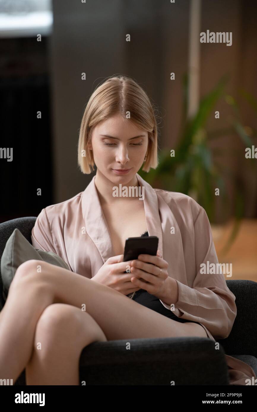 Jeune belle femme assise sur un fauteuil et utilisant son mobile téléphone qu'elle a saisi un message sur le téléphone Banque D'Images