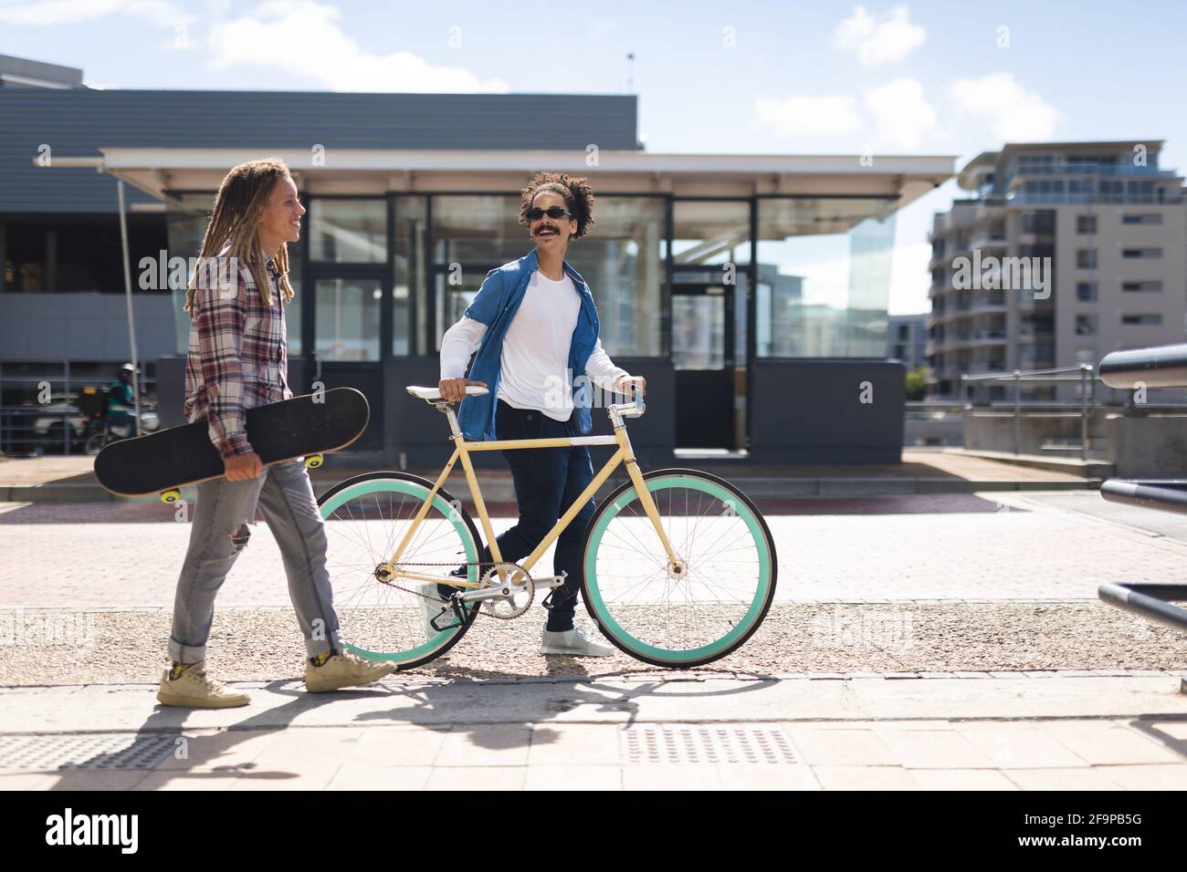 Deux amis de course mixte heureux de sexe masculin à vélo, portant un skateboard dans la rue et parlant. Mode de vie urbain vert, à l'extérieur comme à l'extérieur de la ville. Banque D'Images