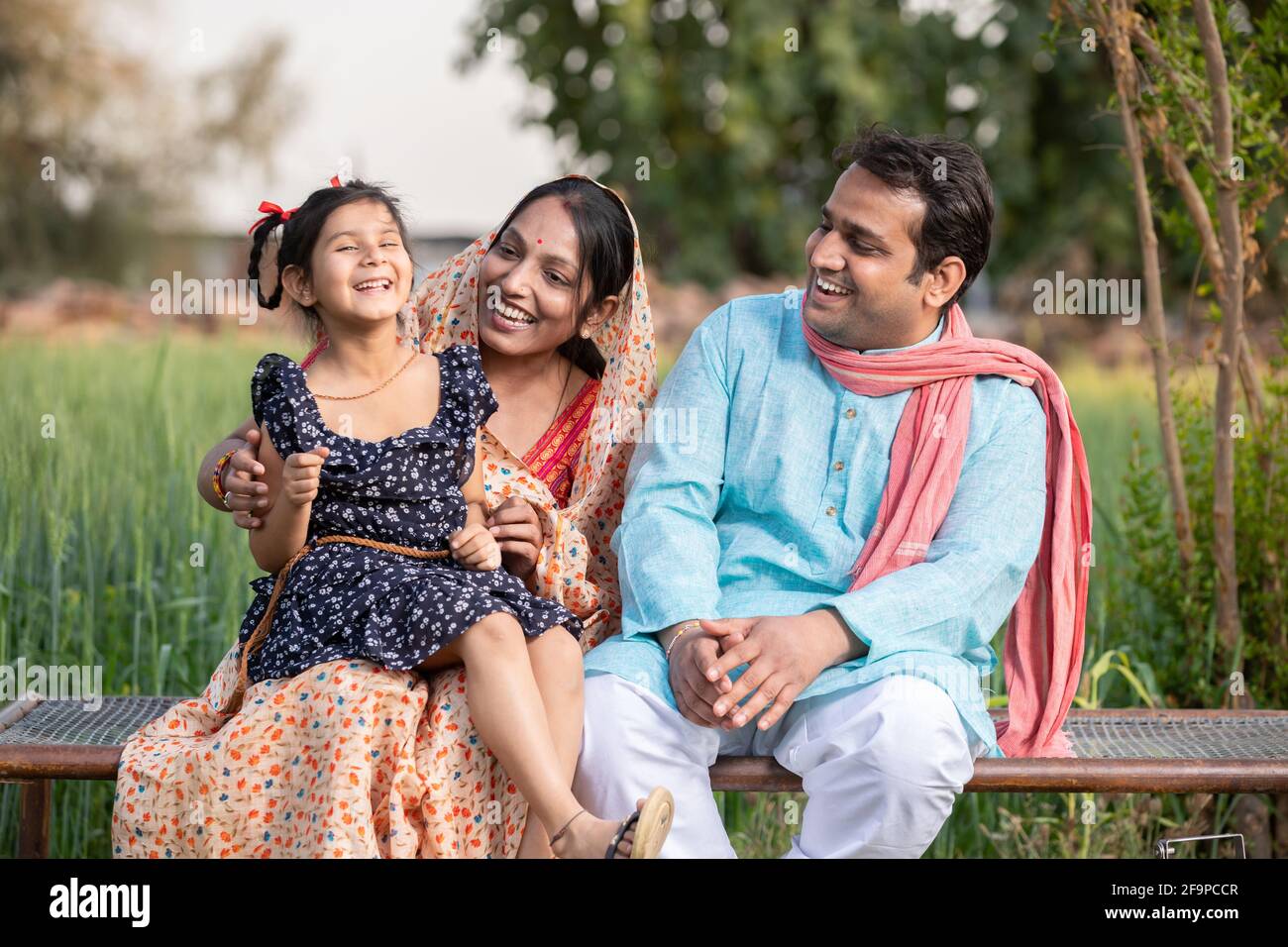 Bonne famille de fermiers indiens assis sur un lit traditionnel au champ agricole dans le village, jeune couple avec leur fille dans la tenue traditionnelle. Banque D'Images