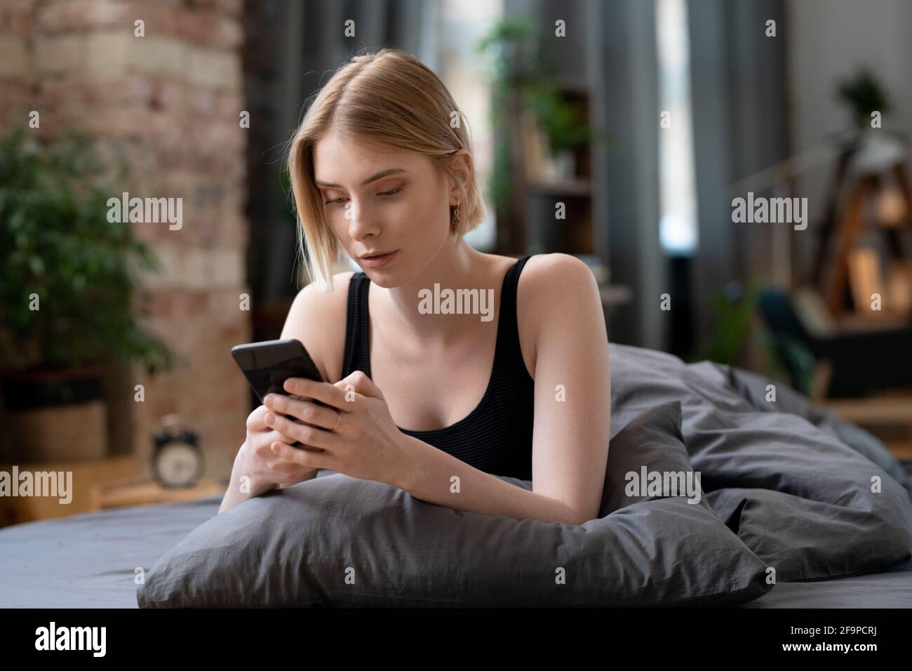 Jeune femme blonde utilisant un téléphone mobile dans son travail en ligne assis au lit le matin Banque D'Images