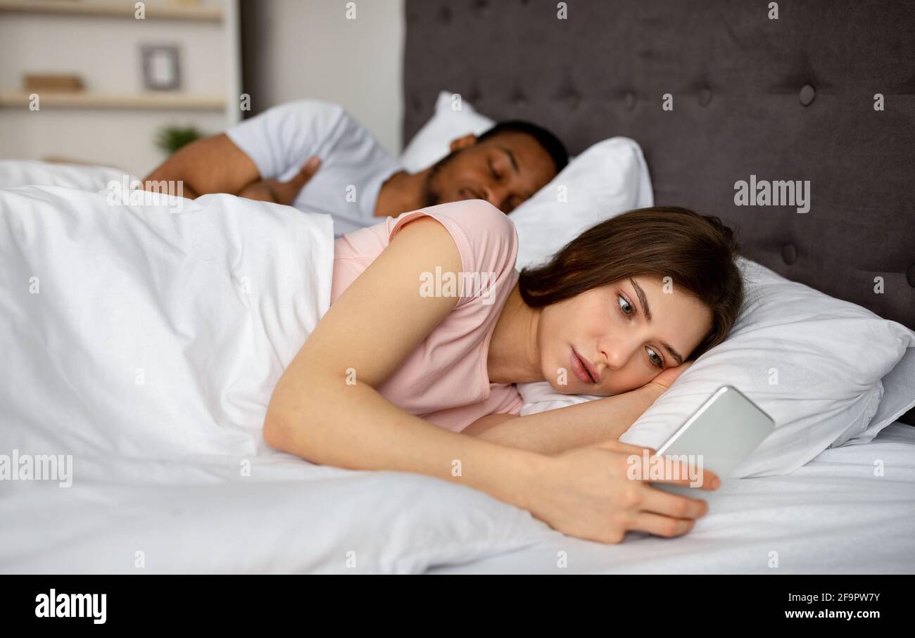 Jeune femme curieuse qui vérifie le smartphone de son mari endormi dans le lit à la maison Banque D'Images