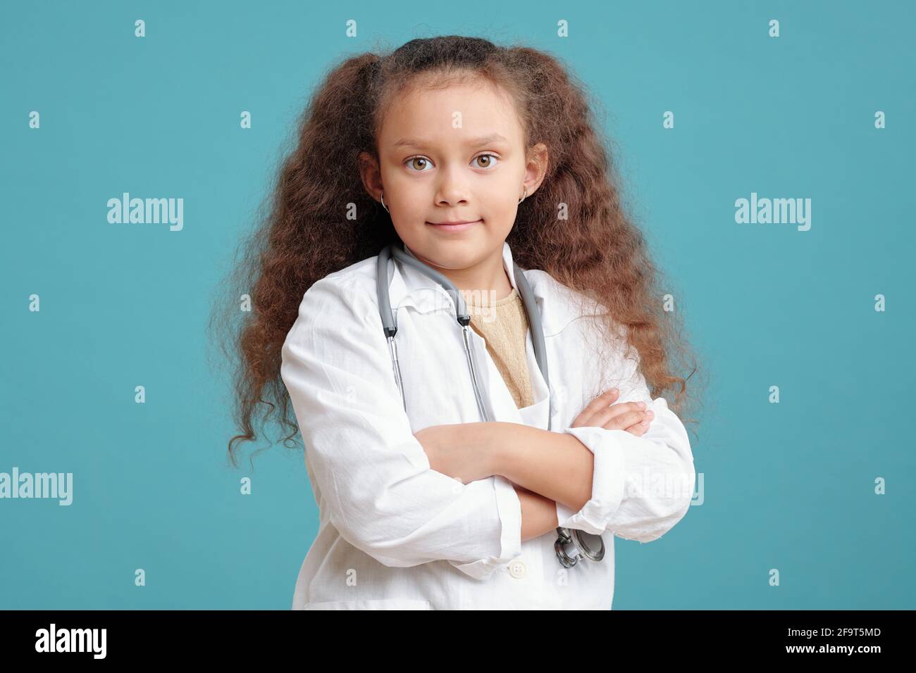 Portrait d'une petite fille aux cheveux bouclés en manteau médical debout, bras croisés et regardant la caméra contre le arrière-plan bleu Banque D'Images