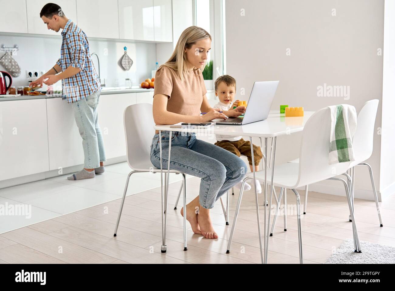 Mère occupée travaillant à la maison en utilisant un ordinateur portable assis à une table avec un enfant mignon fils. Banque D'Images