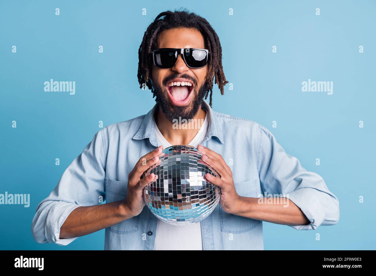 Portrait de gentil gai cool gars tenant dans les mains brillant boule disco ayant l'amusement isolé sur fond bleu clair Banque D'Images