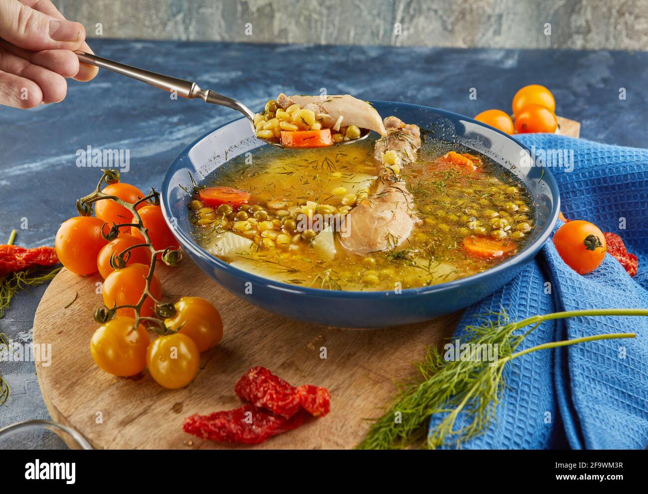 Soupe aux pois avec poulet, carottes, tomates et herbes dans une assiette profonde sur bois avec serviette sur fond bleu. Banque D'Images