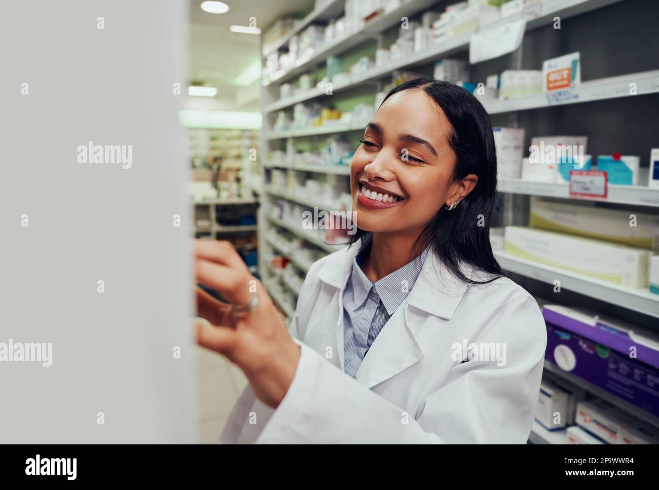 Une femme gaie portant un blouse de travail à la recherche d'un médicament sur une étagère en pharmacie Banque D'Images