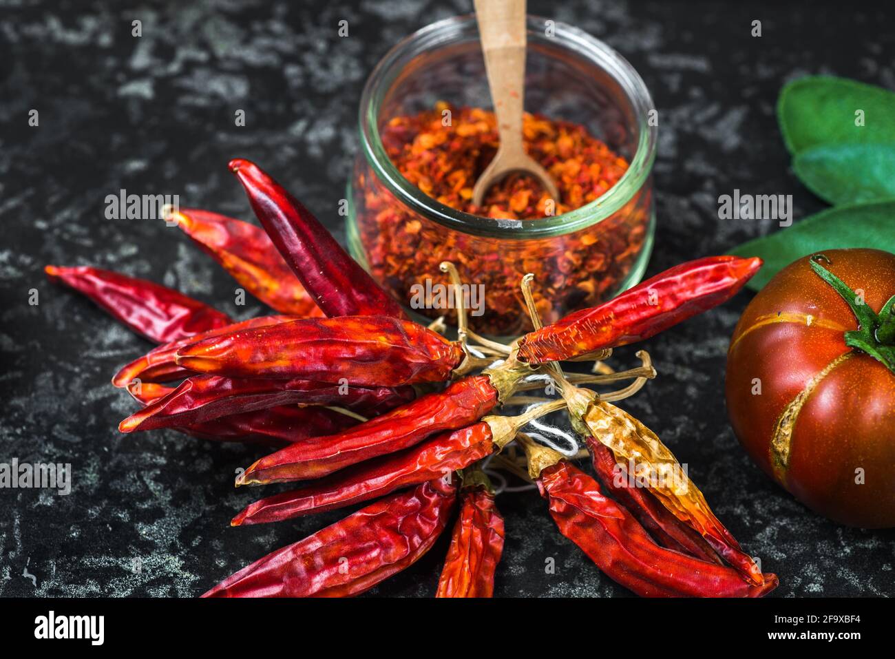 Poivrons rouges chauds séchés en bouquet, condiment chaud au piment écrasé dans un petit bol en verre, tomate et feuille sur fond noir, gros plan. Banque D'Images