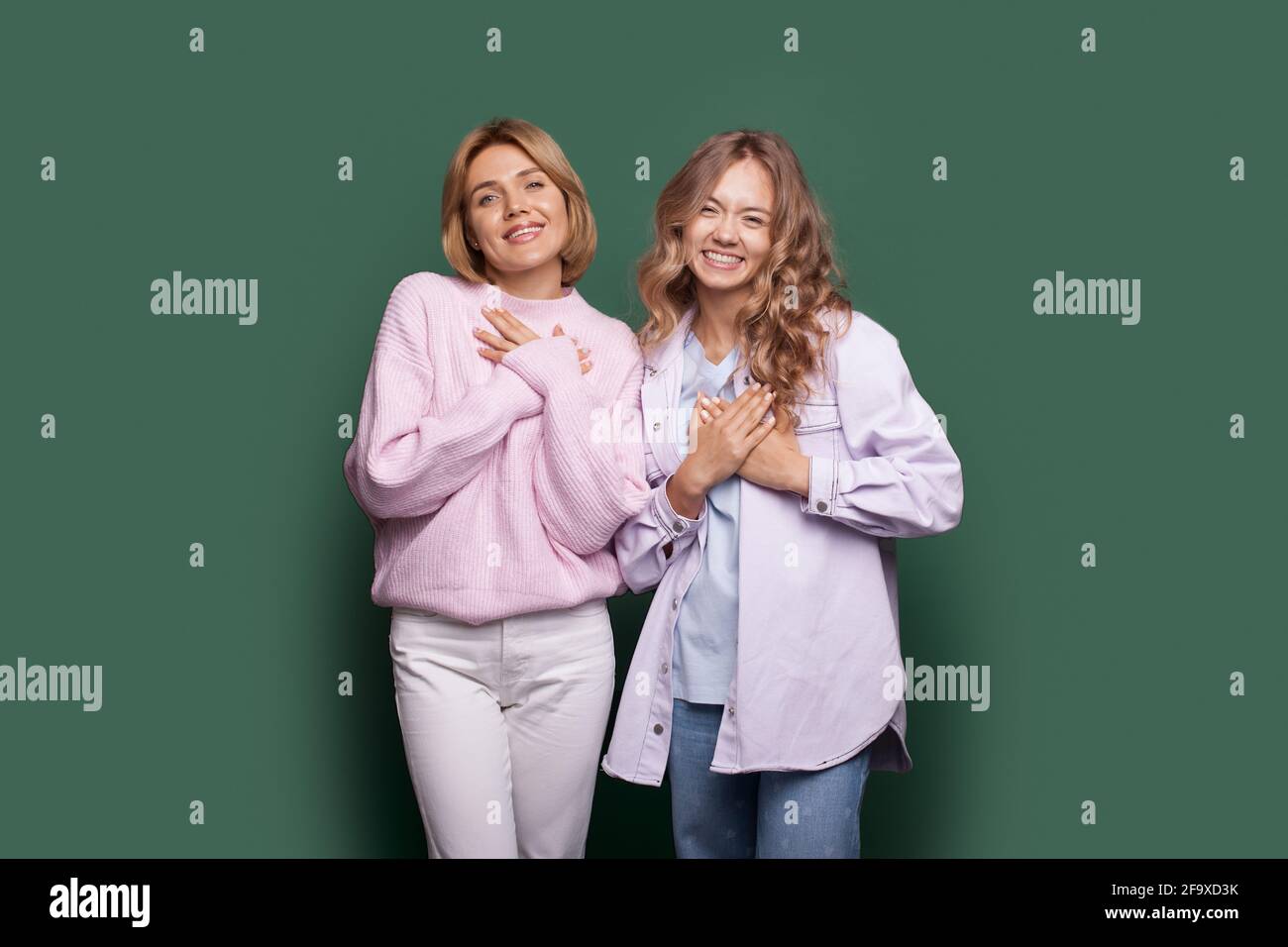 De charmantes sœurs blondes sur un mur vert font le geste signe de coeur touchant leurs coffres Banque D'Images