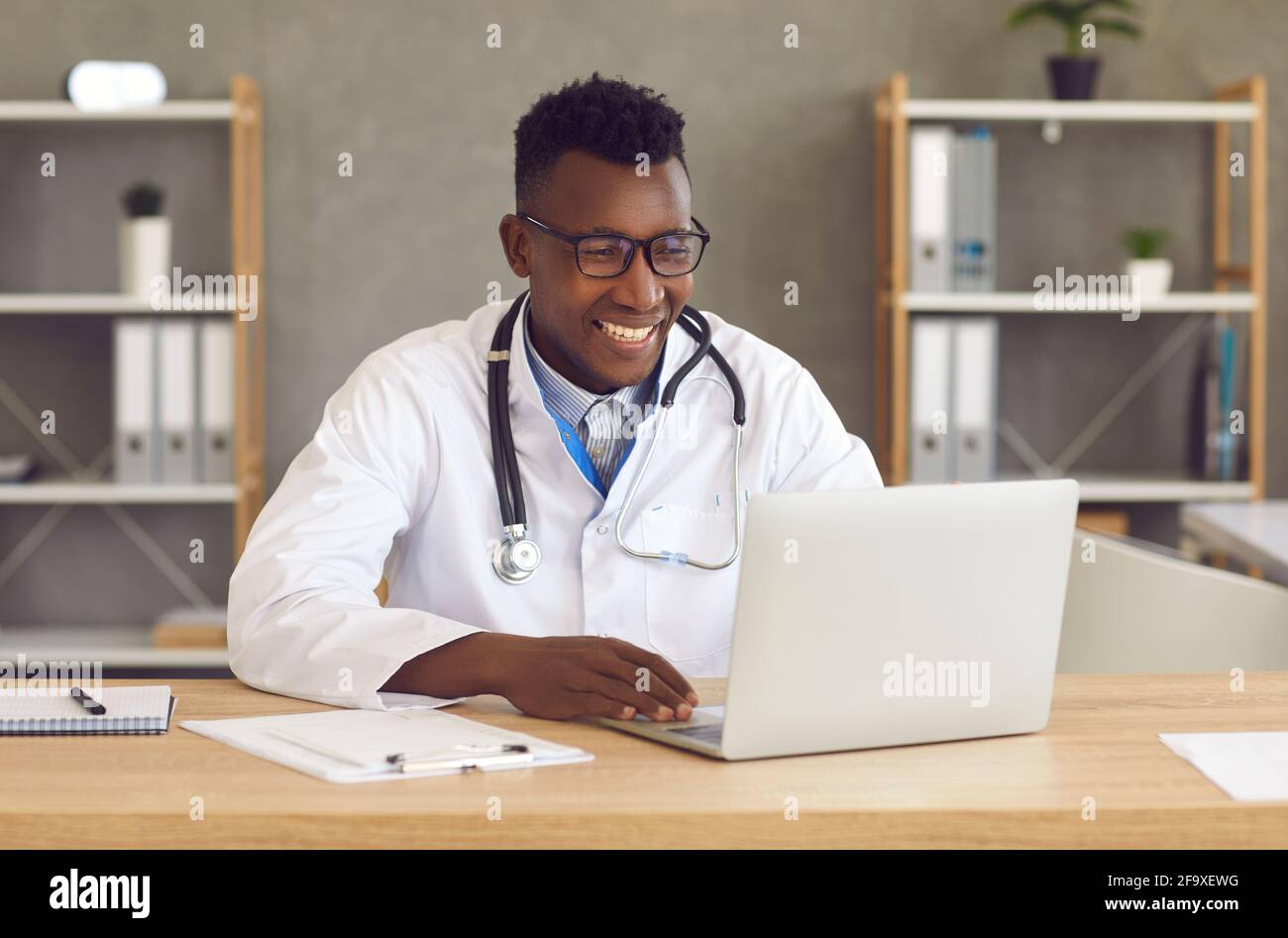 Joyeux médecin noir assis au bureau et utilisant un ordinateur portable pour un appel vidéo avec le patient Banque D'Images Joyeux médecin noir assis au bureau et utilisant un ordinateur portable pour un appel vidéo avec le patient Banque D'Images