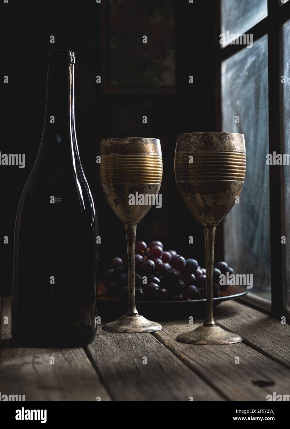 Gobelets et bouteille de vin vintage à côté d'une fenêtre avec raisins en arrière-plan sur une table rustique en bois Banque D'Images
